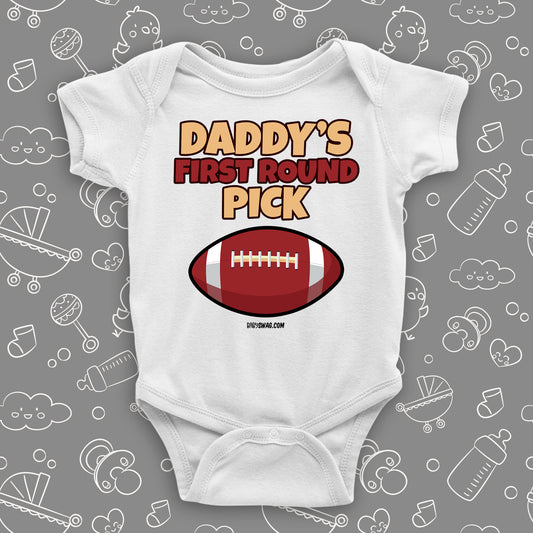 Cool baby onesie saying "Daddy's first round pick" and including the image of a football, color white.