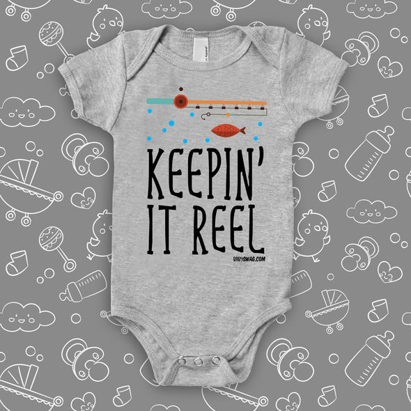 "Keepin' It Reel" written on a grey cute baby onesie, the drawing of a fishing pole and a fish included.