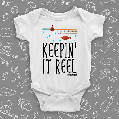 "Keepin' It Reel"  written on a white cute baby onesie, the drawing of a fishing pole and a fish included.