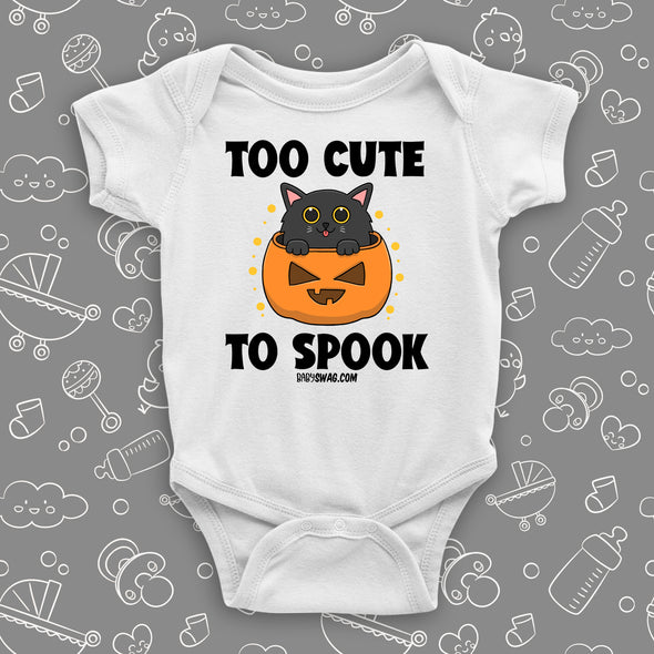 White cute baby onesie saying "Too Cute To Spook" with an image of a kitten coming out of the Halloween pumpkin.